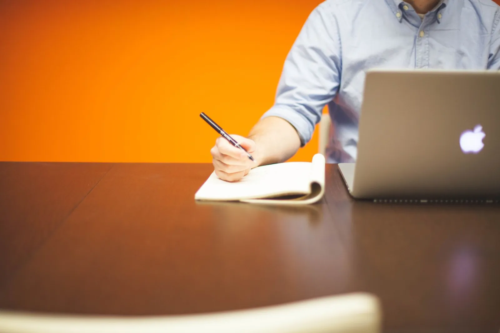 Adult writing notes in a notebook beside a laptop on a wooden desk with orange background.