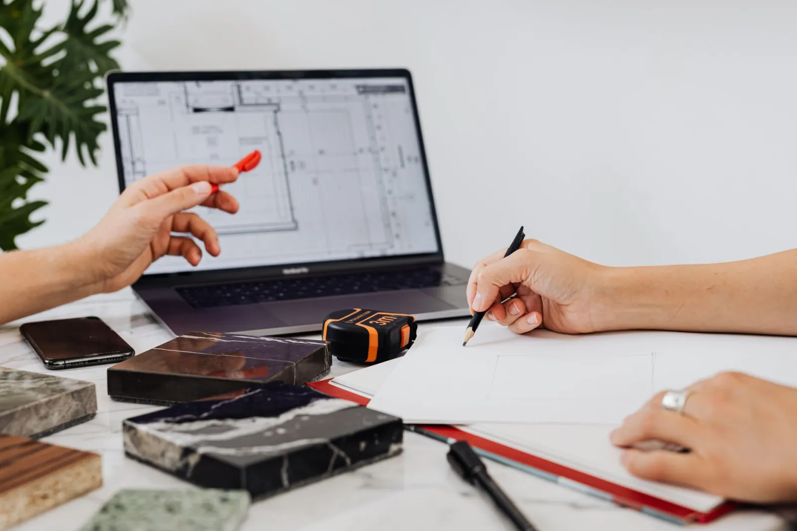 Close-up of hands sketching design plans with tile samples and a laptop, ideal for interior design contexts.