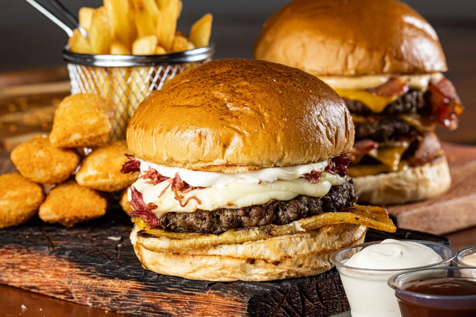 Delicious gourmet burgers with cheese, fries, and dipping sauces on a rustic board.