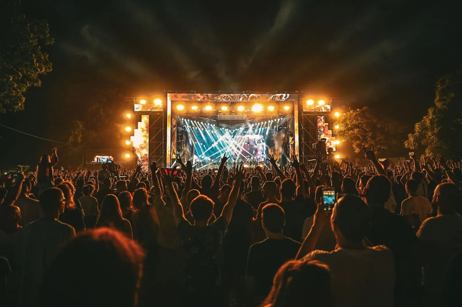 Large crowd enjoying a spectacular live outdoor concert at night with stage lighting effects.