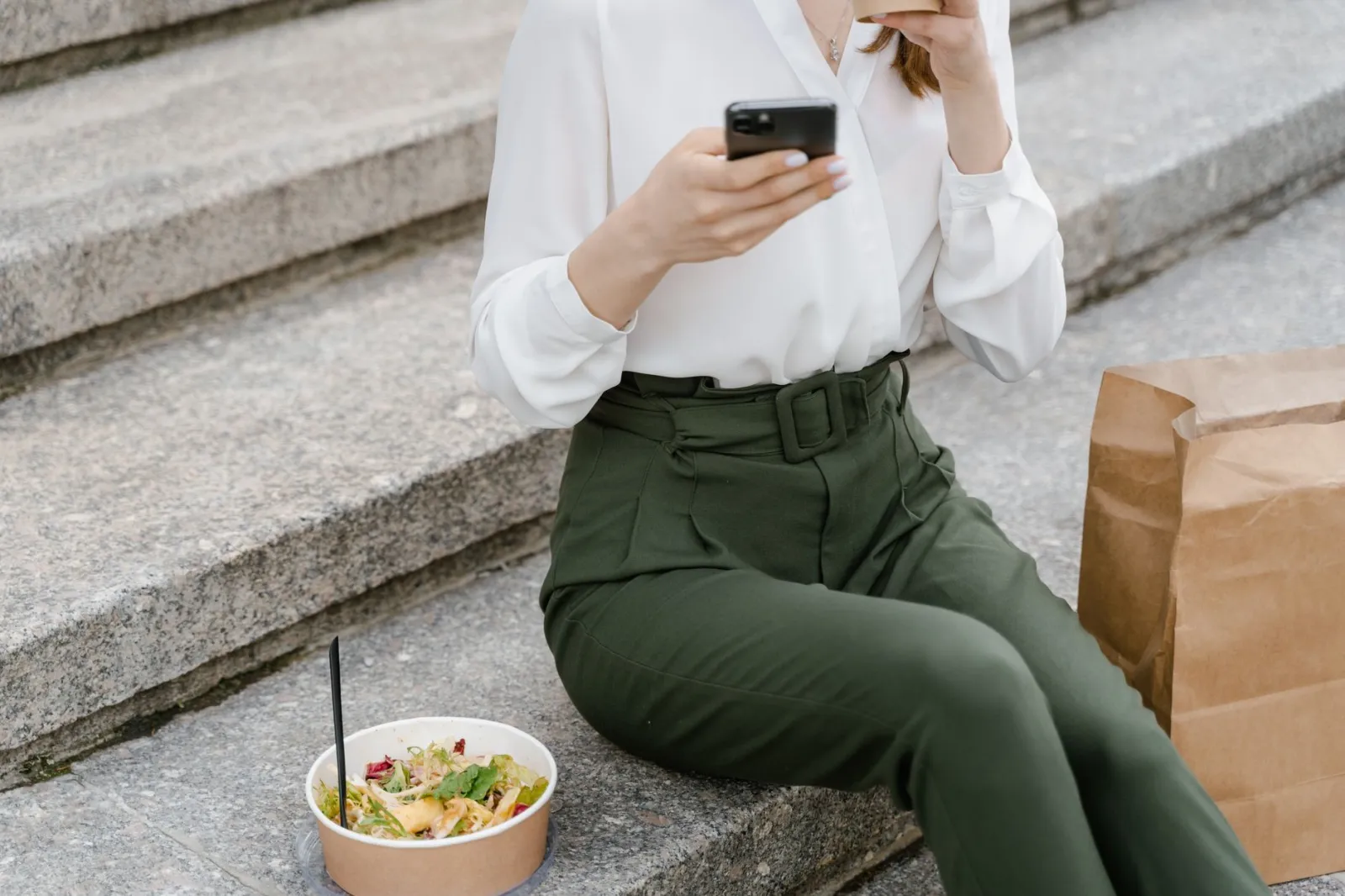 A woman sits on concrete steps with a salad and paper bag, using a smartphone.