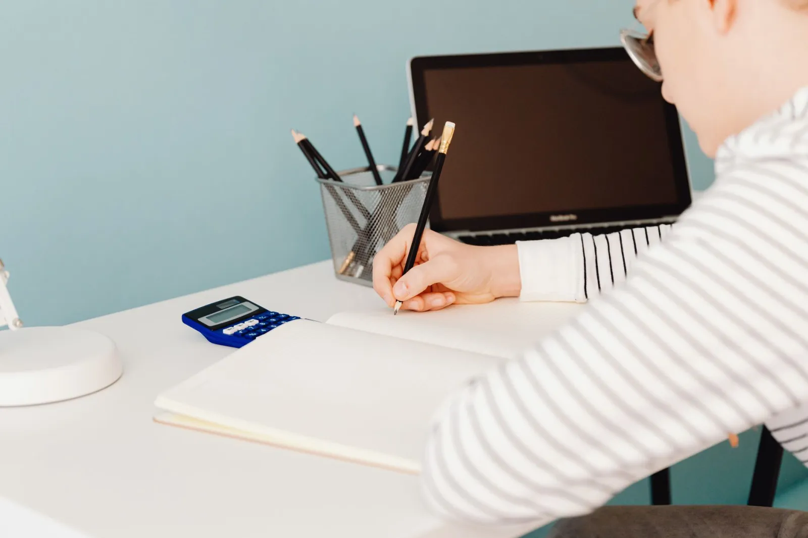 Young adult writing with pencils at a desk with laptop and calculator, perfect for study and work themes.